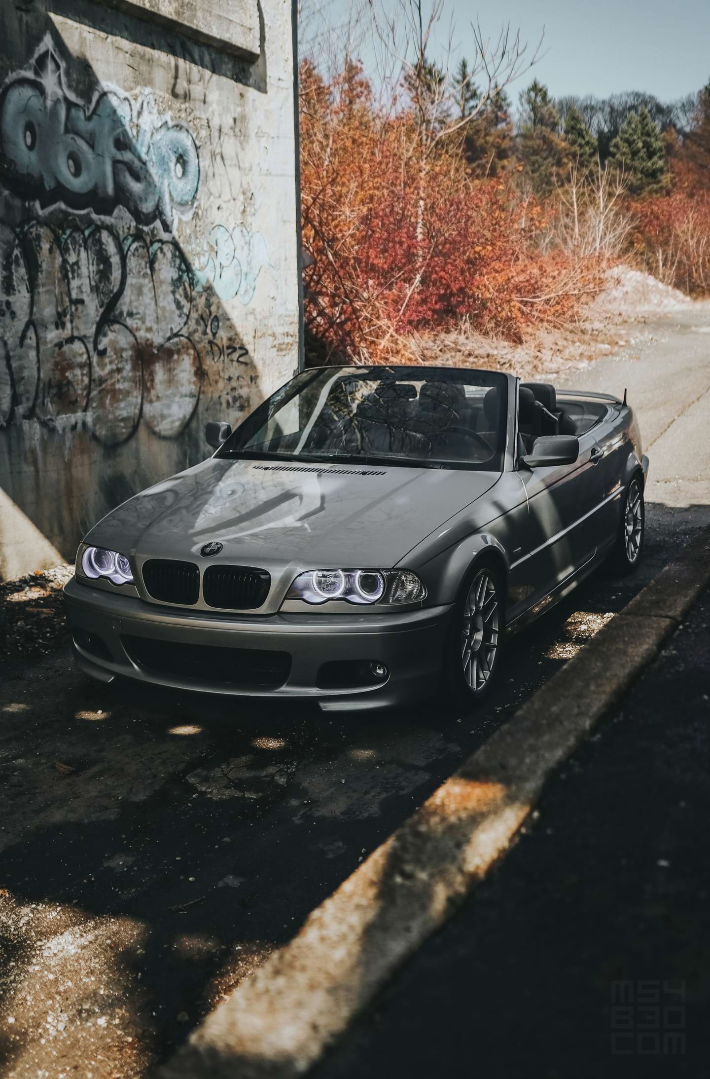 My silver 2002 E46 330Ci parked in shadows under a bridge with fall colors in the background.
