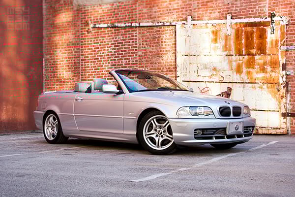 The E46 parked with the top down in front of a red and orange, sunlit brick building.