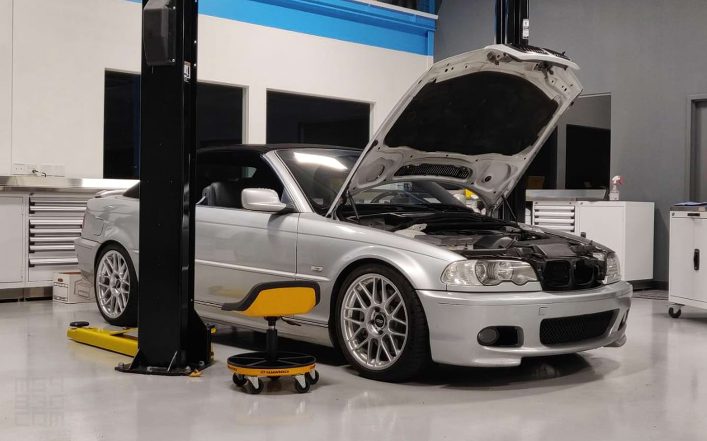 The E46 parked in a professional motorsports garage with the hood open ready for repairs.